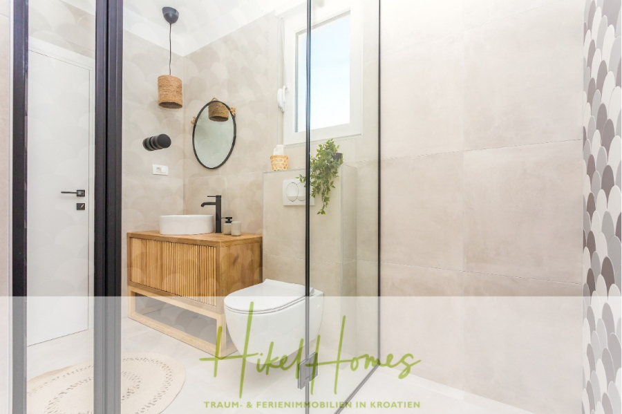 4 Schlafzimmer / 32m² Pool / Sommerküche – Neubauhaus nur 8km von Omis entfernt - Modernes Badezimmer mit hölzernem Waschtisch, rundem Spiegel, Gefäßwaschbecken, wandmontierter Toilette, Glasdusche, Topfpflanze und sanfter natürlicher Beleuchtung. Der Text darüber lautet "Hiket Homes Traum- & Ferienimmobilien in Kroatien. (44129)