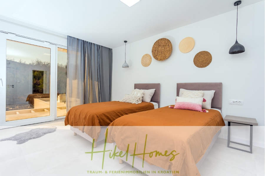 4 Schlafzimmer / 32m² Pool / Sommerküche – Neubauhaus nur 8km von Omis entfernt - Helles, modernes Schlafzimmer mit zwei Einzelbetten in brauner Bettwäsche, dekorativen Kissen, Wandkunst mit Weidenkörben, Pendelleuchten, Glastüren nach draußen und durchsichtigen grauen Vorhängen. "Hike Homes"-Logo am unteren Rand. (44118)