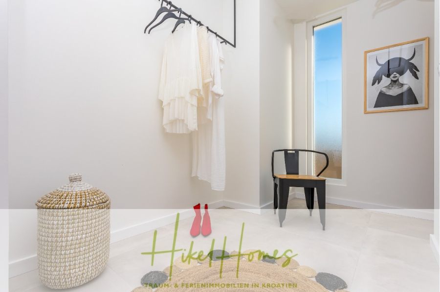 4 Schlafzimmer / 32m² Pool / Sommerküche – Neubauhaus nur 8km von Omis entfernt - Ein minimaler, heller Umkleideraum mit weißen Wänden, einem Hängeregal mit weißer Kleidung, einem geflochtenen Korb, einem schwarzen Stuhl, einem hohen Fenster und gerahmter minimalistischer Kunst an der Wand. (44125)