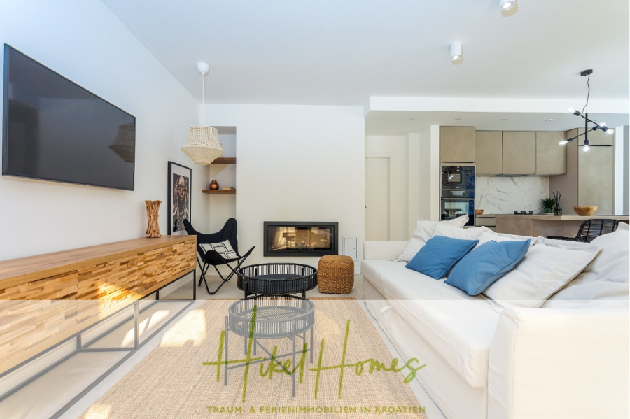 4 Schlafzimmer / 32m² Pool / Sommerküche – Neubauhaus nur 8km von Omis entfernt - Helles, modernes Wohnzimmer mit weißem Sofa, blauen Kissen, geflochtenen Stühlen, an der Wand montiertem Fernseher, Kamin, runden Couchtischen und offener Küche im Hintergrund. Das Logo "Hikel Homes" überlagert den Boden. (44113)