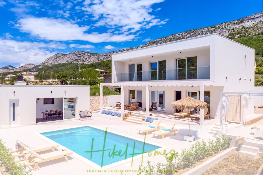 4 Schlafzimmer / 32m² Pool / Sommerküche – Neubauhaus nur 8km von Omis entfernt - Moderne weiße Villa mit einem Pool, Sonnenliegen und einer schattigen Terrasse vor grünen Hügeln und felsigen Bergen unter einem strahlend blauen Himmel. Ess- und Sitzbereiche im Freien sind sichtbar; das Branding lautet "Hikel Homes". (44130)