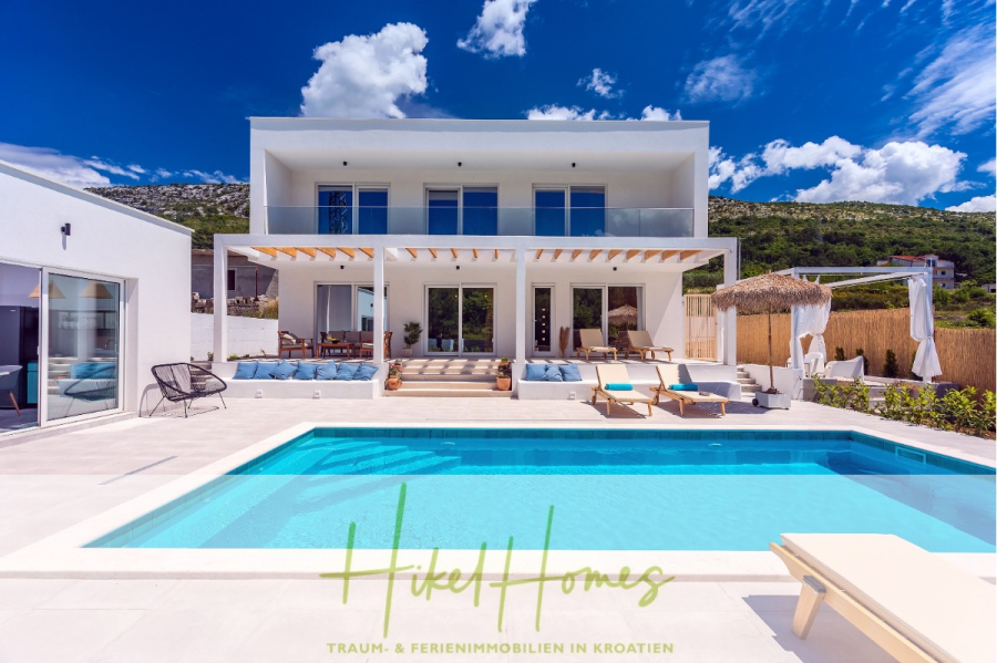 4 Schlafzimmer / 32m² Pool / Sommerküche – Neubauhaus nur 8km von Omis entfernt - Moderne weiße Villa mit Balkonen und großen Fenstern, einem Außenpool, Sonnenliegen, einer schattigen Terrasse und Hügeln im Hintergrund unter einem blauen Himmel. Der Text am unteren Rand lautet "Hikel Homes-Traum- & Ferienimmobilien in Kroatien. (44102)