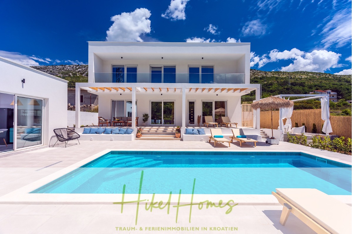 Moderne weiße Villa mit Balkonen und großen Fenstern, einem Außenpool, Sonnenliegen, einer schattigen Terrasse und Hügeln im Hintergrund unter einem blauen Himmel. Der Text am unteren Rand lautet "Hikel Homes-Traum- & Ferienimmobilien in Kroatien. - 4 Schlafzimmer / 32m² Pool / Sommerküche – Neubauhaus nur 8km von Omis entfernt