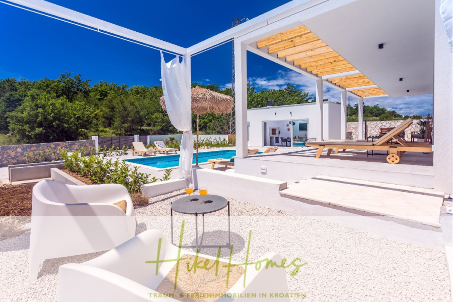 4 Schlafzimmer / 32m² Pool / Sommerküche – Neubauhaus nur 8km von Omis entfernt - Eine moderne Außenterrasse mit weißen Möbeln, einem kleinen Tisch mit zwei Getränken, Liegestühlen am Swimmingpool und einer modernen weißen Villa, umgeben von Grün unter blauem Himmel. (44109)