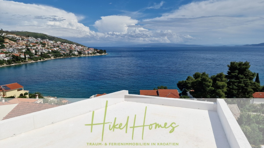 Luxus Meerblick 77m² Ensemble mit 2 Schlafzimmern, Terrasse und Südausrichtung (1. OG) - Blick auf eine Küstenstadt mit Häusern in Hanglage mit Blick auf ein ruhiges blaues Meer unter einem teilweise bewölkten Himmel. Im Vordergrund sind ein weißes Dach und ein Glasgeländer zu sehen. Der Text am unteren Rand lautet "Hikel Homes Traum- & Ferienimmobilien in Kroatien". (46834)